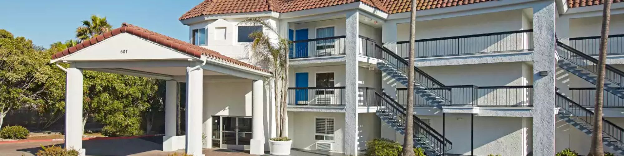 An exterior view of a two-story building with a red-tiled roof, palm trees, and outdoor staircases, likely a hotel or motel, during a sunny day.