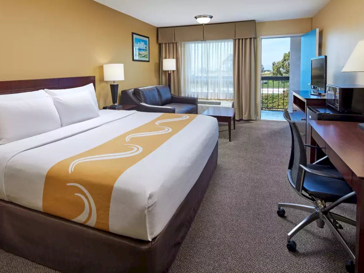 This image shows a hotel room with a king-size bed, desk, chair, TV, sofa, coffee table, bedside tables with lamps, window, and patio door.