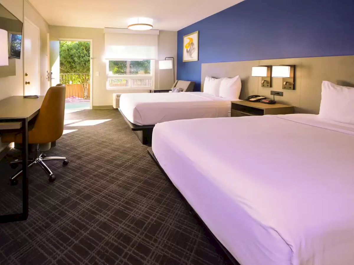 The image shows a clean, modern hotel room with two double beds, a desk with a chair, a flat-screen TV, and a window with a view outside.