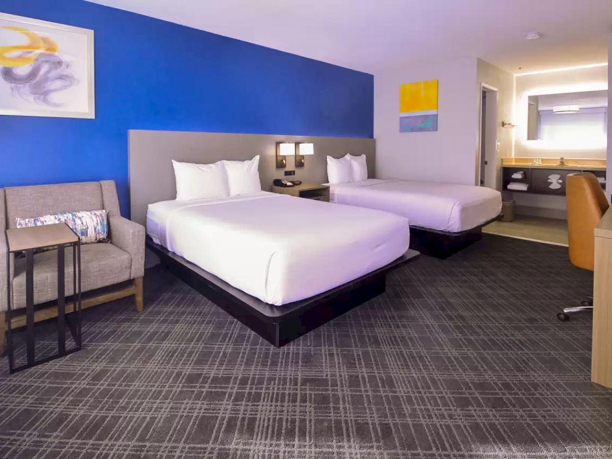 The image shows a modern hotel room with two double beds, a chair with a side table, a desk with a chair, and colorful wall art.