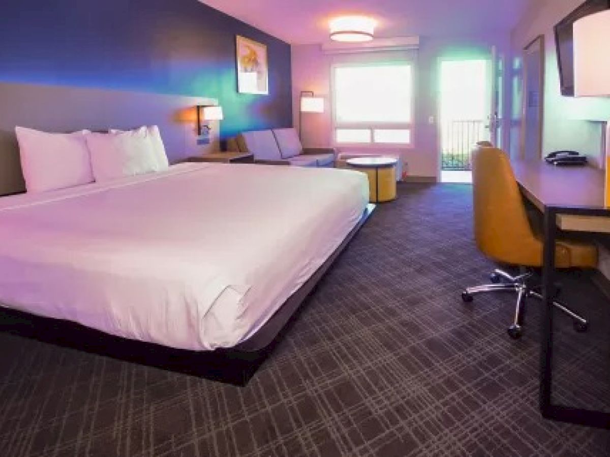 A modern hotel room features a large bed, a sofa, a desk with a chair, and a balcony with a view. Warm lighting and decor create a welcoming ambiance.