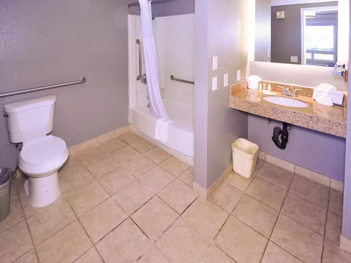 The image shows a clean bathroom with a toilet, bathtub with a curtain, sink with a mirror, and a waste bin on tiled flooring.