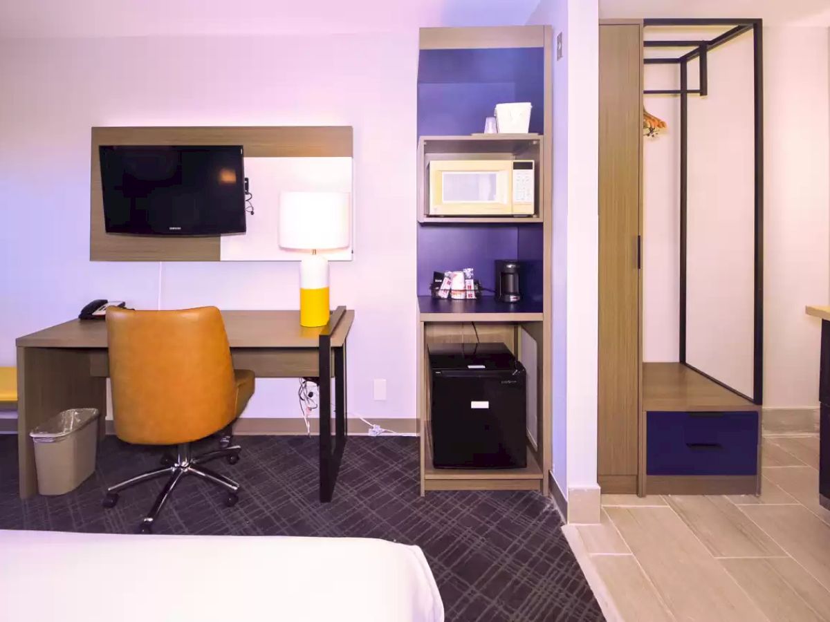 A hotel room features a desk, chair, TV, lamp, microwave, coffee maker, mini-fridge, closet, and neatly arranged towels, all well-lit.