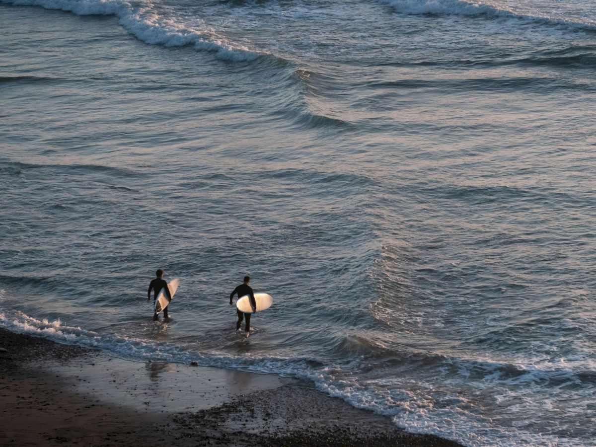 Two surfers carrying surfboards are walking into the ocean, seemingly preparing for a surf session as gentle waves approach the shoreline.