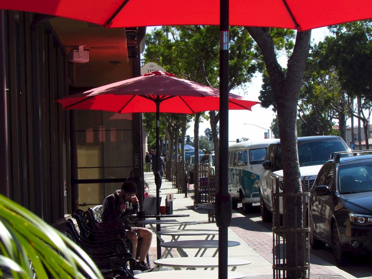 Sidewalk café with red umbrellas and outdoor seating beside parked cars on a sunny day. Trees and buildings line the street in the background.