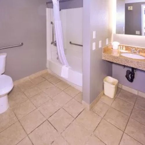 A clean bathroom with a toilet, bathtub, shower curtain, vanity mirror, sink, countertop, and a small trash can.