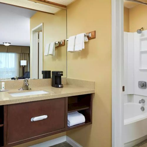 A bathroom with a sink, mirror, coffee maker, towels, toiletries, and a connected room with a tub, shower, toilet, and curtained window in the background.