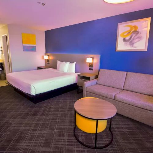 A modern hotel room features a bed, couch, desk, and bright decor with a view into the bathroom area, blending comfort with contemporary style.