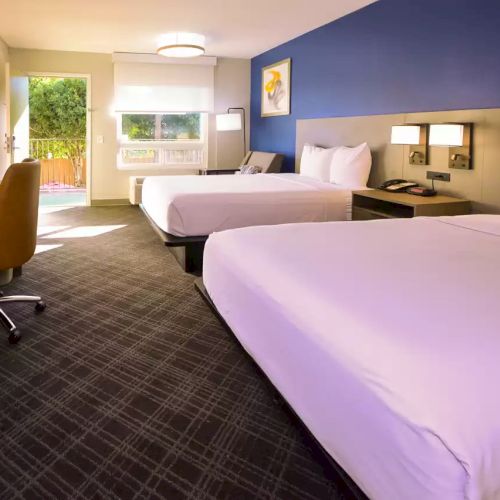 The image shows a hotel room with two double beds, a desk with a chair, modern decor, and a door leading to an outdoor area.