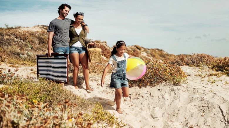 A smiling family with beach accessories is walking through sand dunes toward the beach, enjoying a sunny day.