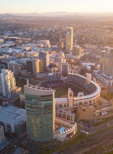 An aerial view of a cityscape at sunset, featuring a central stadium surrounded by high-rise buildings and busy streets, with mountains in the distance.