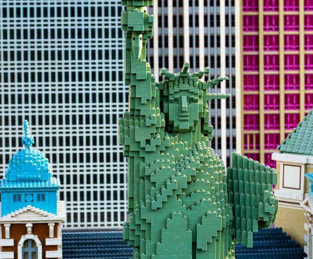 The image shows a LEGO model of the Statue of Liberty against a backdrop of colorful LEGO buildings.